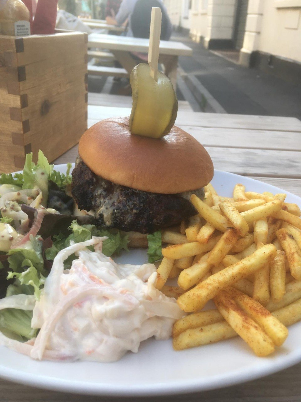 A burger topped with a gherkin on a cocktail stick, served with seasoned fries, salad and coleslaw at one of the pub's outdoor picnic tables