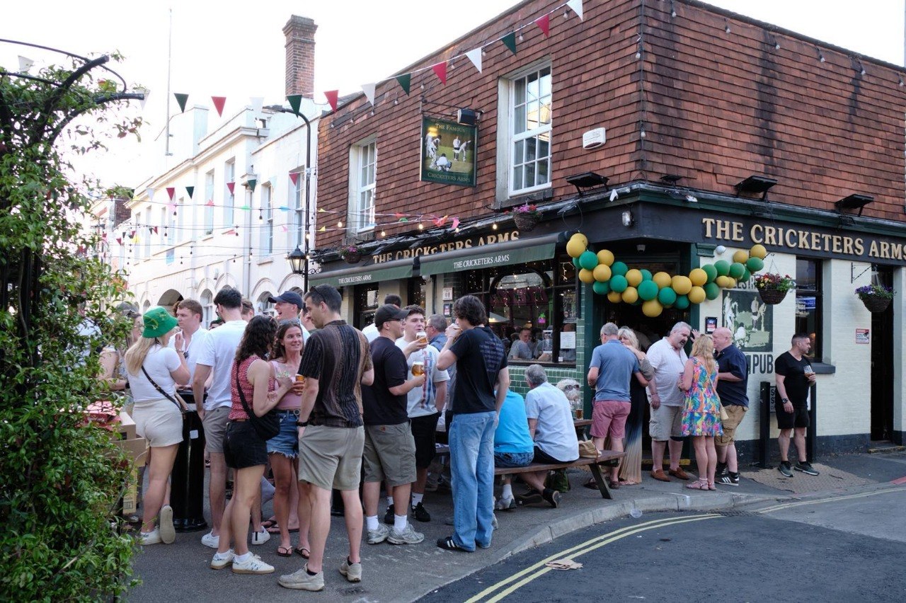 A crowd filling the pavement outside The Cricketers Arms during a street event, with bunting strung overhead and a green and gold balloon arch around the pub sign