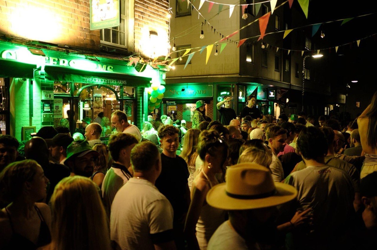 A dense crowd outside The Cricketers Arms at night, lit green, with colourful bunting overhead and the pub's illuminated signage above the doorway