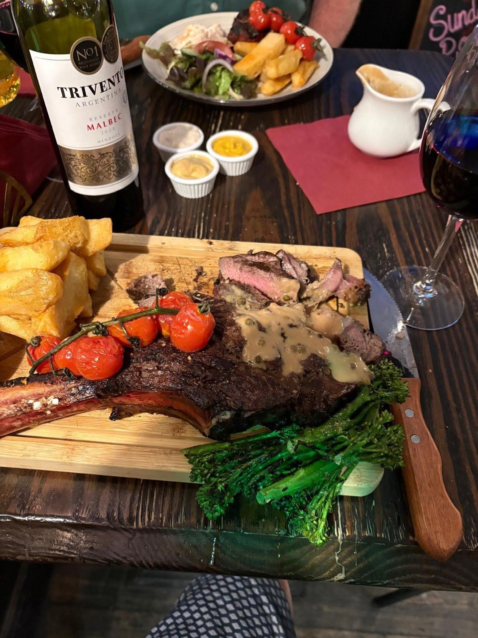 A tomahawk steak on a wooden board with peppercorn sauce, chunky chips, vine tomatoes and tenderstem broccoli, served with red wine on a pub table